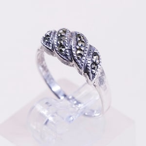May include: A silver-toned ring with a braided design. The ring features a series of small, dark gemstones set along the braided pattern. The ring is displayed on a clear stand against a white background, showcasing its intricate details.