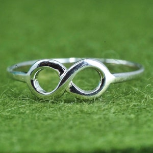 May include: A silver-colored ring featuring an infinity symbol design. The ring is set against a green background, highlighting the polished metal. The infinity symbol is a continuous loop, symbolizing eternity.