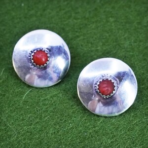 May include: A pair of silver stud earrings, each featuring a central red coral stone. The earrings have a circular design with a textured border. The background is a solid green colour.