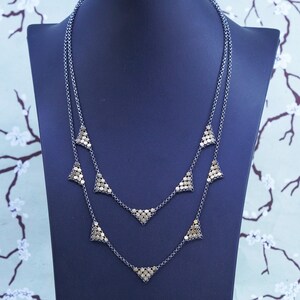 May include: A layered necklace with a dark silver chain and multiple triangle pendants. Each pendant is made of small, round, gold-colored beads. The necklace is displayed on a dark blue jewelry stand against a floral background.