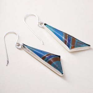 May include: A pair of silver earrings with a triangular design. The earrings feature a mosaic of blue, turquoise, and brown stones set in a silver frame. The earrings have a hook closure.