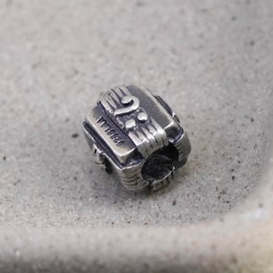 Puede incluir: Un pequeño charm plateado con un diseño de nota musical. El charm presenta una clave de sol y notas musicales, con el texto "0238LA" visible. El charm tiene forma cilíndrica con grabados detallados.