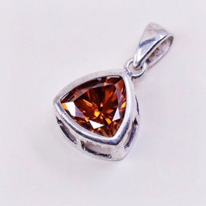 May include: A silver pendant with a triangular, faceted, orange gemstone. The pendant has a simple, openwork design around the stone and a loop for a chain. The gemstone is a warm, amber color, catching the light.