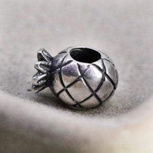 Puede incluir: Un charm de color plateado con forma de piña. El charm tiene una superficie texturizada con un patrón de diamantes y una pequeña abertura en la parte superior. La corona de la piña está detallada con varias hojas.