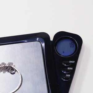 May include: A silver necklace with a textured circular pendant is placed on a digital scale. The scale displays a weight of 1.8 grams. The scale is black with a silver weighing surface and a digital display.