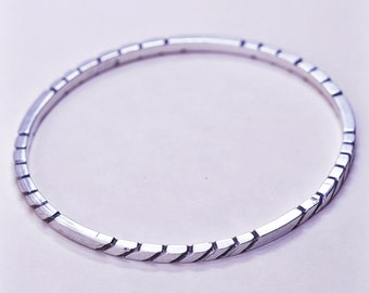 May include: A silver-toned bangle bracelet with a geometric design. The bracelet features a series of rectangular segments with diagonal cuts, creating a textured appearance. The bracelet is circular and appears to be made of metal.