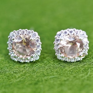 May include: A pair of stud earrings with a central peach-colored stone surrounded by a halo of smaller, clear stones. The earrings are set against a green background, showcasing their sparkle and design.