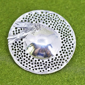 May include: A silver-coloured brooch shaped like a hat. The brooch has a central, rounded design with a textured surface, encircled by a perforated brim. A decorative bow and leaves are attached to the hat's side. The brooch is set against a green background.