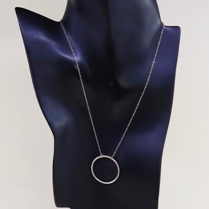 May include: A silver-toned necklace with a delicate chain and a textured, open circle pendant. The necklace is displayed on a black mannequin, showcasing its simple elegance. The pendant's diameter is approximately 1 inch.