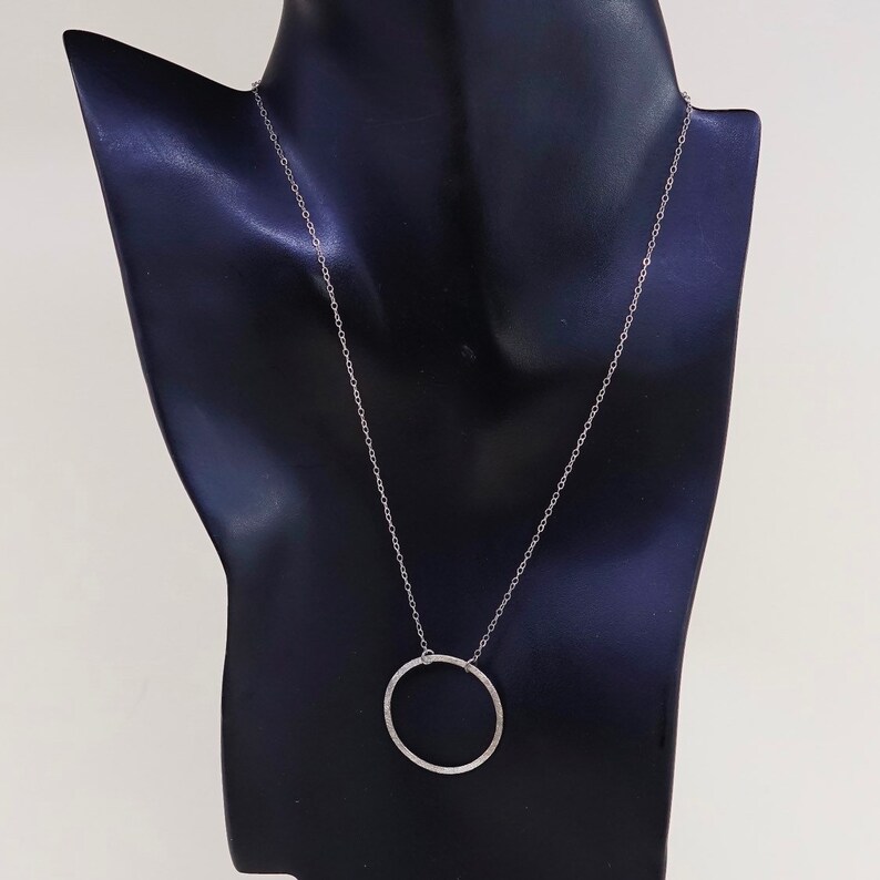 May include: A silver-toned necklace with a delicate chain and a textured, open circle pendant. The necklace is displayed on a black mannequin, showcasing its simple elegance. The pendant's diameter is approximately 1 inch.