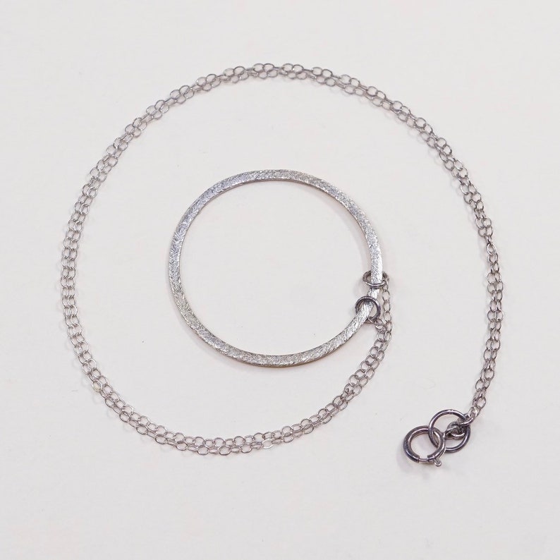 May include: A silver necklace featuring a textured, open circle pendant. The pendant is connected to a delicate chain with a lobster clasp. The necklace is displayed on a white background.