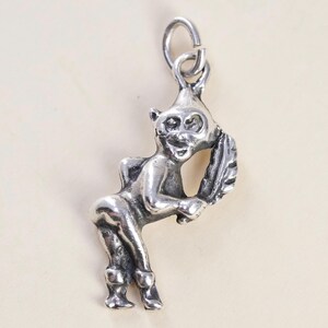 May include: A silver-toned metal charm shaped like a small, standing imp or sprite figure. The figure has pointed ears, a smiling expression, and is holding a large feather-like object. A circular jump ring is attached to the top for hanging.