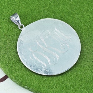 May include: A silver-colored pendant with a circular design. The pendant features an engraved monogram of the letters "N" and "M" in a cursive style. The pendant has a small loop at the top for a chain.