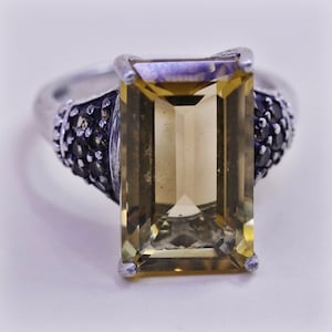May include: A silver ring featuring a large, rectangular, light-yellow gemstone. The gemstone is the focal point, set in a silver band with black accent stones on each side of the gemstone.