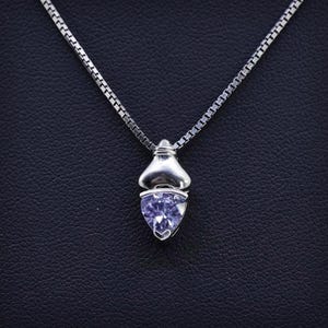 May include: A silver necklace with a pendant featuring a light purple, heart-shaped gemstone. The pendant is set in a silver metal casing and hangs from a delicate silver chain. The necklace is displayed against a dark background.