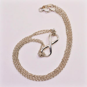 May include: A silver-toned necklace with an infinity symbol pendant. The chain is made of multiple strands of small, linked circles. The pendant is a smooth, polished infinity shape. The necklace is displayed on a light pink background.