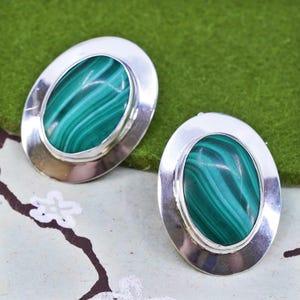 May include: A pair of oval-shaped earrings with a polished silver frame. Each earring features a vibrant green malachite stone with distinctive swirling patterns. The earrings are displayed on a green and white patterned surface.