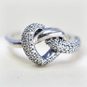 May include: A silver-toned ring featuring a central knot design. One half of the knot is smooth and polished, while the other half is encrusted with numerous small, clear, sparkling stones that cover the surface of the metal band.