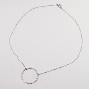 May include: A silver-toned necklace featuring a delicate chain and a large, textured open circle pendant. The pendant is connected to the chain with small, dark-colored rings. The necklace is displayed against a white background.