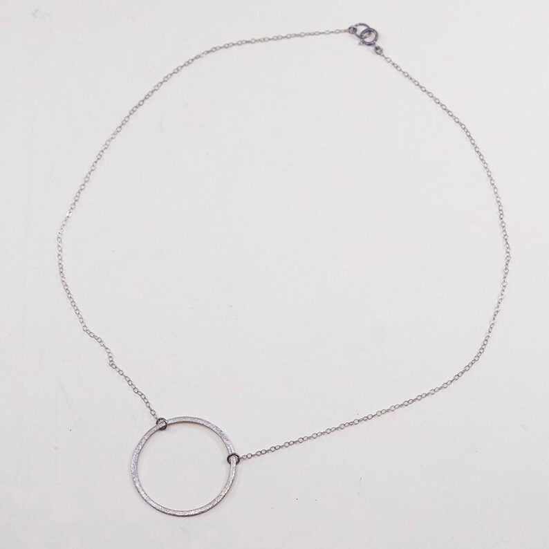 May include: A silver-toned necklace featuring a delicate chain and a large, textured open circle pendant. The pendant is connected to the chain with small, dark-colored rings. The necklace is displayed against a white background.