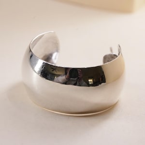 May include: A polished silver cuff bracelet with a rounded, open design. The bracelet reflects the surrounding environment, showcasing a smooth, shiny surface. This bold piece of jewelry is a statement accessory.
