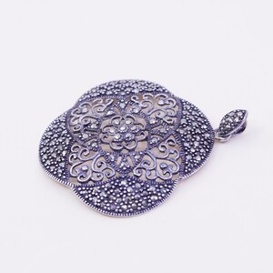May include: A silver-toned pendant with a floral design. The pendant has an openwork pattern with small, round, dark stones. The pendant is in the shape of a four-leaf clover. The bail is a small, rounded loop.