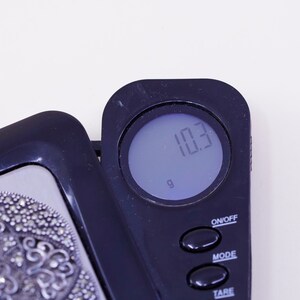 May include: A digital scale displays a weight of 10.3 grams. The scale is black with a digital display and several buttons. A decorative silver object is partially visible on the left side of the image.