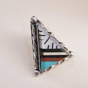 May include: A silver-toned triangular ring with a colorful inlaid design. The inlay features a white section with black lines, and bands of black, white, red, and turquoise. The ring has a braided border.