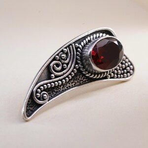Könnte beinhalten: Silberfarbener Ring mit einem ovalen, tiefroten Edelstein. Der Ring weist aufwendige, wirbelnde Muster und eine strukturierte Oberfläche auf. Der Edelstein ist in einer Zargenfassung gefasst, und der Ring hat ein gebogenes, asymmetrisches Design.