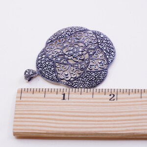 May include: A detailed, silver-toned pendant with an openwork floral design. The pendant is approximately 2 inches in diameter and features a small loop for a chain. The surface is covered in small, sparkling accents, creating a vintage aesthetic.