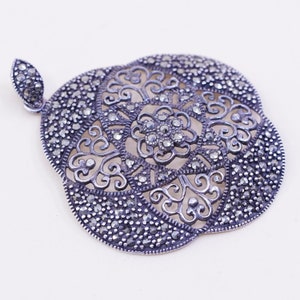 May include: A silver-toned pendant with a floral design. The pendant features an openwork pattern with a central flower and surrounding scrollwork, accented with small, dark stones. The bail is a small, leaf-shaped design.