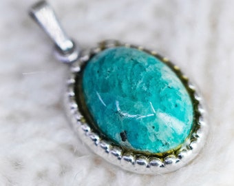 May include: A silver pendant with an oval, turquoise-colored stone. The stone has a mottled appearance with variations in color. The pendant has a decorative, beaded border and a silver bail for a chain.