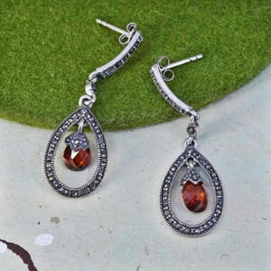 May include: A pair of silver-toned drop earrings. Each earring features a teardrop-shaped frame with small, dark accents surrounding a red-brown gemstone. The earrings have a post-back closure and a curved top.