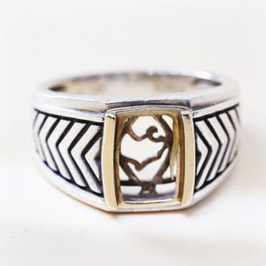 May include: A silver ring featuring a central rectangular gold-toned frame surrounding an openwork design of intertwined branches. The band displays a repeating chevron pattern in oxidised silver, creating a high-contrast look across the metal surface.