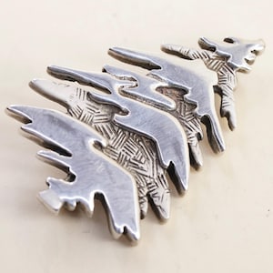 May include: A silver-toned metal brooch shaped like a stylized evergreen tree. The design features layered, horizontal branches with a smooth, polished finish on the edges and a textured, cross-hatched pattern on the recessed sections.