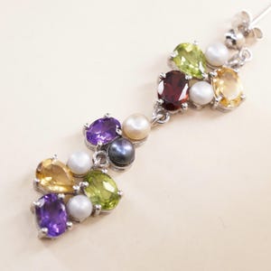 Peut inclure: Boucle d'oreille en argent ornée d'un ensemble de pierres précieuses colorées et de perles. La boucle d'oreille présente des pierres d'améthyste, de citrine, de péridot et de grenat de taille ovale, ainsi que des perles blanches et grises foncées.