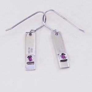 May include: A pair of silver-colored dangle earrings. Each earring features a rectangular bar with a small, round, pink gemstone set at the bottom. The earrings have a simple, elegant design with a hook closure.