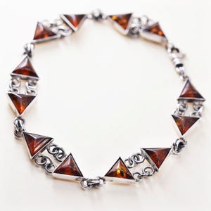 May include: A silver bracelet with triangular amber stones. The amber stones are set in silver frames and linked by silver chains. The bracelet is shown against a white backdrop, highlighting its design and craftsmanship.
