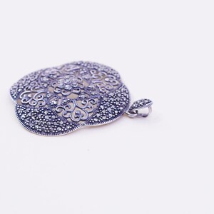 May include: A silver-toned pendant with an intricate, openwork floral design. The pendant is encrusted with small, sparkling stones, and has a bail for a chain. The overall shape is a stylized flower, perfect for adding a touch of elegance.