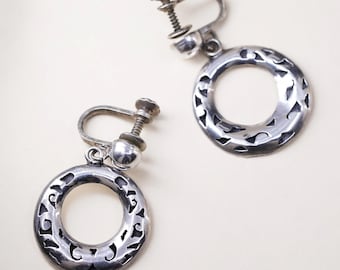 May include: A pair of silver-toned screw-back earrings. Each earring features a circular design with an open center and decorative cut-out patterns around the edge. The earrings are displayed against a light background.