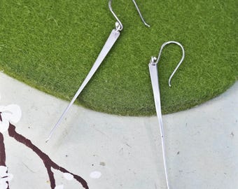 Può includere: Un paio di orecchini argentati dal design minimalista. Ogni orecchino presenta una lunga e sottile barra rettangolare che si assottiglia fino a un punto, sospesa da un gancio curvo. Gli orecchini sono esposti su uno sfondo verde e bianco.