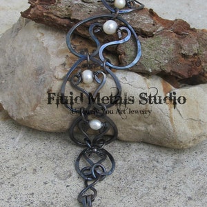 Peut inclure: Un bracelet en métal noir avec un design délicat et complexe présentant de multiples boucles et volutes. Le bracelet est orné de quatre perles blanches.