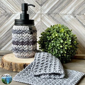 May include: A grey and white crocheted soap dispenser bottle with a black pump. The bottle is sitting on a wooden surface with a grey crocheted washcloth next to it. A small green bush is in the background.
