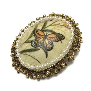 May include: Oval-shaped brooch with a butterfly design. The brooch features a central image of an orange and black butterfly on a light green background, surrounded by faux pearls and a gold-toned floral frame.