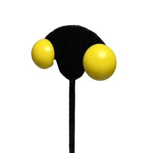 May include: A pair of bright yellow, round clip-on earrings. The earrings are a solid, glossy yellow color and are displayed on a black velvet earring stand. The earrings have a simple, classic design.