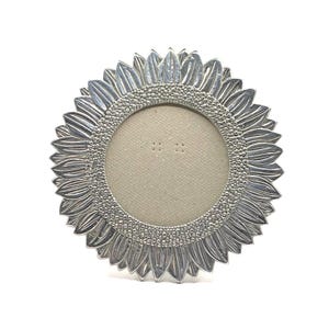 May include: A circular silver-toned picture frame featuring a sunflower-inspired design. The outer edge consists of sculpted metal petals, while the inner border surrounding the beige circular insert is decorated with a textured, beaded pattern.