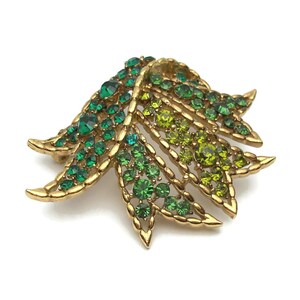 May include: A gold-toned leaf-shaped brooch with a gradient of green rhinestones. The brooch features a detailed design with varying shades of green stones, from deep emerald to light chartreuse, set in a gold metal frame.