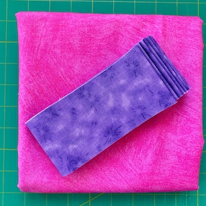 May include: A bright pink fabric square with a textured pattern, topped with a folded purple fabric rectangle. The purple fabric has a speckled design. The items are on a green cutting mat.