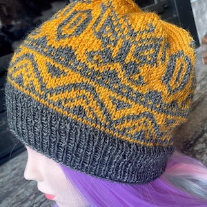 May include: A knitted beanie hat in shades of mustard yellow and gray. The hat features a geometric pattern across the crown and a solid gray ribbed brim. The hat is displayed on a mannequin head with purple hair.