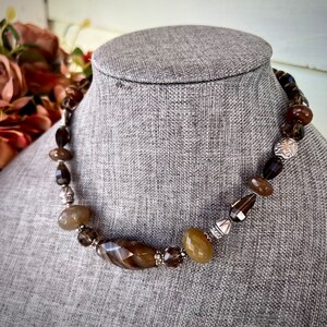 May include: A brown and beige agate gemstone necklace with silver accents. The necklace is made with faceted and smooth beads.